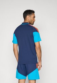 Tmavomodré polokošeľa s krátkymi tyrkysovými rukávmi, s červenými a žltými pruhmi pozdĺž ramien, doprevádzaná zladenými šortkami.