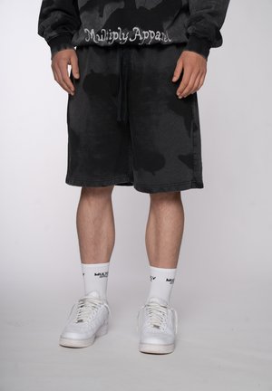 Mann trägt schwarze Batik-Shorts, schwarzen Pullover mit „Multiply Apparel“, weiße Marken-Socken und weiße Sneakers, steht vor einfachem Hintergrund.