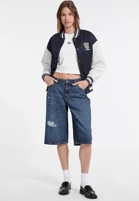 Marineblauwe en grijze varsityjas met witte strepen, gecombineerd met blauwe denim shorts met distressed accenten. Model draagt zwarte loafers en witte sokken.