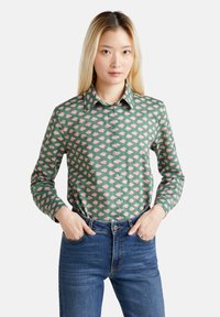 Camicia verde a maniche lunghe con pattern floreale rosa, colletto, abbinata a jeans blu con una vestibilità aderente.