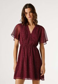 Vestido burdeos con mangas cortas y acampanadas. Hecho de tela ligera, cuenta con escote en v y lunares blancos, ajustado en la cintura.