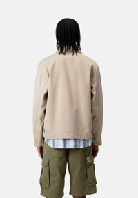 Chaqueta ligera beige de corte recto, con tela texturizada y un diseño sencillo, combinada con una camisa de rayas azul y shorts cargo en color oliva.