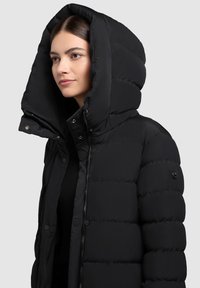 Černá prošívaná bunda s velkou kapucí, zapínáním na zip a patentními knoflíky. Má hladkou texturu a kulatý logo detail na rukávu.