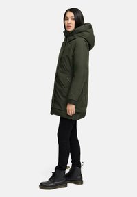 OLivengrøn parka-jakke med hætte, der har lynlås, to sidelommer og elastiske manchetter, parret med sorte skinny jeans og støvler.