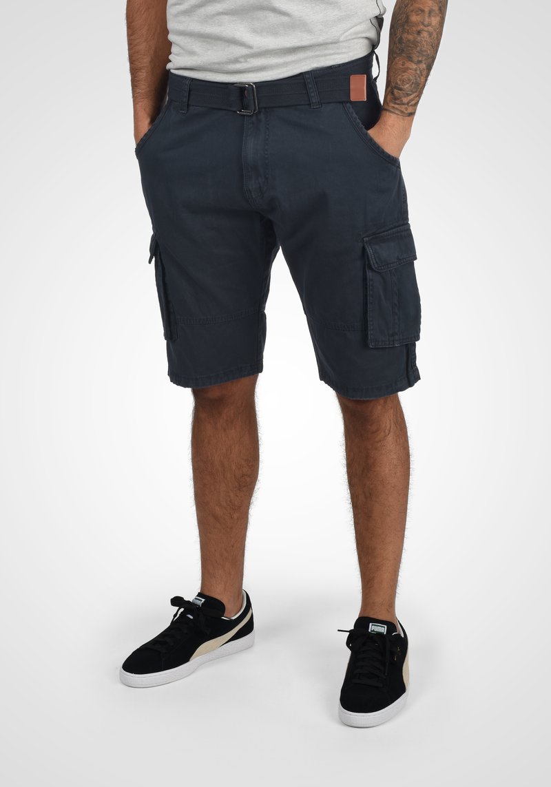 Marineblaue Cargo-Shorts mit mehreren Taschen, einem Gürtel und einem lässigen Schnitt, kombiniert mit schwarzen Sneakers mit einer weißen Sohle und beigen Akzenten.