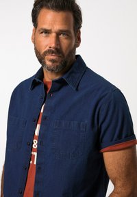 Camicetta blu navy a maniche corte in tessuto denim con chiusura a bottoni e due tasche sul petto. Accento rosso sulla manica e t-shirt a girocollo sottostante.