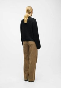 Černý texturovaný svetr s dlouhými rukávy, skombinovaný s hnědými sametovými kalhotami s vysokým pasem a širokými nohavicemi, které mají žebrovaný vzor a zadní kapsy.
