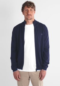 Veste zippée bleu marine avec un col haut, coupe slim, poignets côtelés, et accents bleu clair. Portée sur un t-shirt blanc. Fond uni.