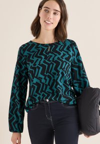 Multifarvet top med et abstrakt mønster i teal og sort, rund halsudskæring med en nøglehulldetalje, parret med mørke jeans.