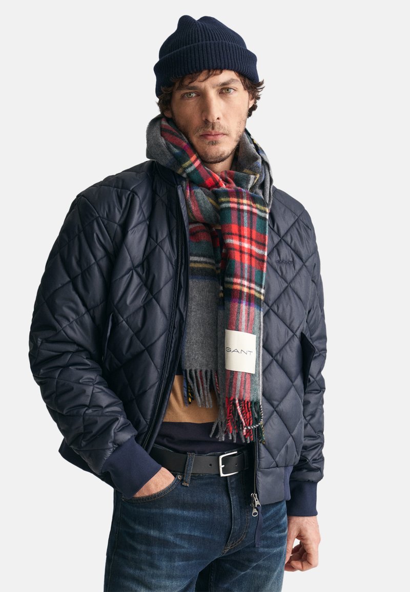Námořnická prošívaná bomber bunda, šedá mikina viditelná, červený a zelený kostkovaný šál, tmavé džíny, černý opasek, neformální zimní outfit.