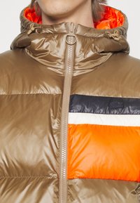 Personne portant une doudoune brillante marron avec des panneaux bicolores orange, blanc et noir et une capuche doublée orange, zippée.