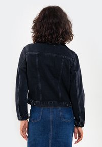 Musta farkkutakki, jossa on istuva malli, näkyvät tikkaustyöt, napit yksityiskohdissa ja klassinen kaulus, pidetty sinisen farkkuhameen päällä.