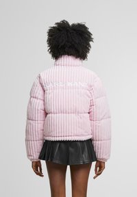 Giacca puffer in corduroy rosa con righe verticali, colletto alto e logo ricamato "KARL KANI" sulla parte superiore della schiena. Una gonna nera completa il look.