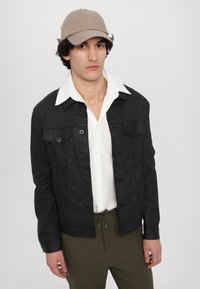 Svart denimjakke med knapper, to brystlommer og en glatt tekstur. Kombinert med en hvit skjorte og olivengrønne bukser.