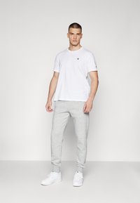 Witte T-shirt met klein logo op de borst; grijze sweatpants met geribde boorden; witte sneakers; effen achtergrond.