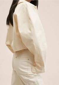 Kremowa krótka kurtka wykonana z lekkiego materiału, z zapięciem na guziki, widocznymi szwami i luźnym krojem. Noszona z pasującymi spodniami.