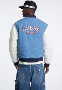 Kurtka bombka dżinsowa w jasnoniebieskim kolorze, z białymi skórzanymi rękawami, granatowymi ściągaczami i haftem "GUESS JEANS" na plecach.