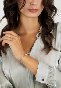 Bracciale rigido in argento con due estremità texturizzate, caratterizzato da piccoli accenti scintillanti. Indossato sopra una blusa leggera e lucente con colletto a punta.