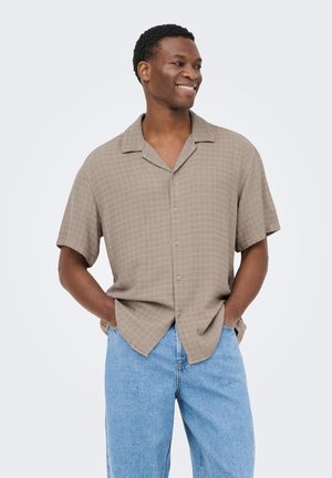 Hombre sonriente con las manos en los bolsillos, lleva una camisa beige de manga corta abotonada y vaqueros azules claros, mirando hacia un lado contra un fondo liso.