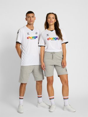 Mann und Frau stehen nebeneinander und tragen weiße "Verändere alles"-Shirts, graue Shorts, weiße Turnschuhe und regenbogenfarbene gestreifte Socken.