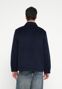 Jūras zila vilnas jaka ar klasisku apkakli un garām piedurknēm, ar pogotu aproci un gludu tekstūru. Attēlots no aizmugures.