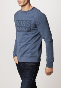 Sudadera azul hecha de una tela suave con un cuello redondo, que presenta el texto "BOSS ORANGE" en un tono más oscuro en la parte frontal.