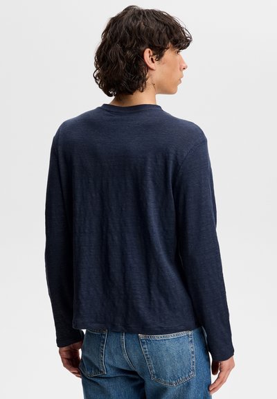Homme aux cheveux bouclés portant une chemise à manches longues bleu marine et un jean bleu, de dos sur un fond uni gris clair.
