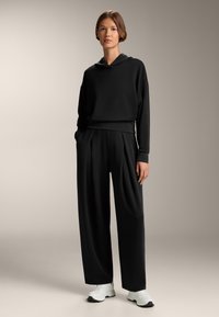 Felpa nera aderente con tasca frontale e cappuccio, abbinata a pantaloni neri a vita alta e gamba larga con pieghe. Sneaker bianche.