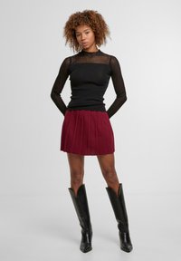 Černý žebrovaný top s dlouhým rukávem a vysokým výstřihem, zkombinovaný s bordó plisovanou minisukní a černými koženými vysokými botami s špičkou.