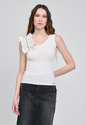 Jonge vrouw met lang golvend haar, gekleed in een witte mouwloze top met ruches en strassdetail op de schouder, en een zwarte denimrok.