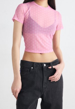Młoda kobieta w różowym krótkim t-shircie z małymi powtarzającymi się logo oraz czarnych, wysokich jeansach, z jedną ręką w kieszeni.