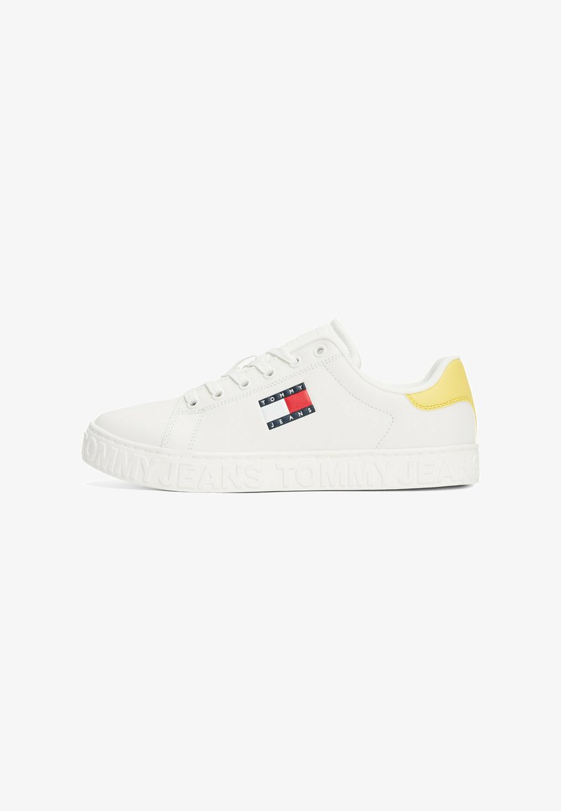 Baskets blanches en matériau synthétique texturé. Elles présentent un accent jaune au niveau du talon, ainsi qu'un patch logo avec un design bleu, rouge et blanc.