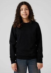 Joven con cabello rizado, vistiendo un sudadero negro y jeans azules, de pie frente a un fondo liso de color gris claro.
