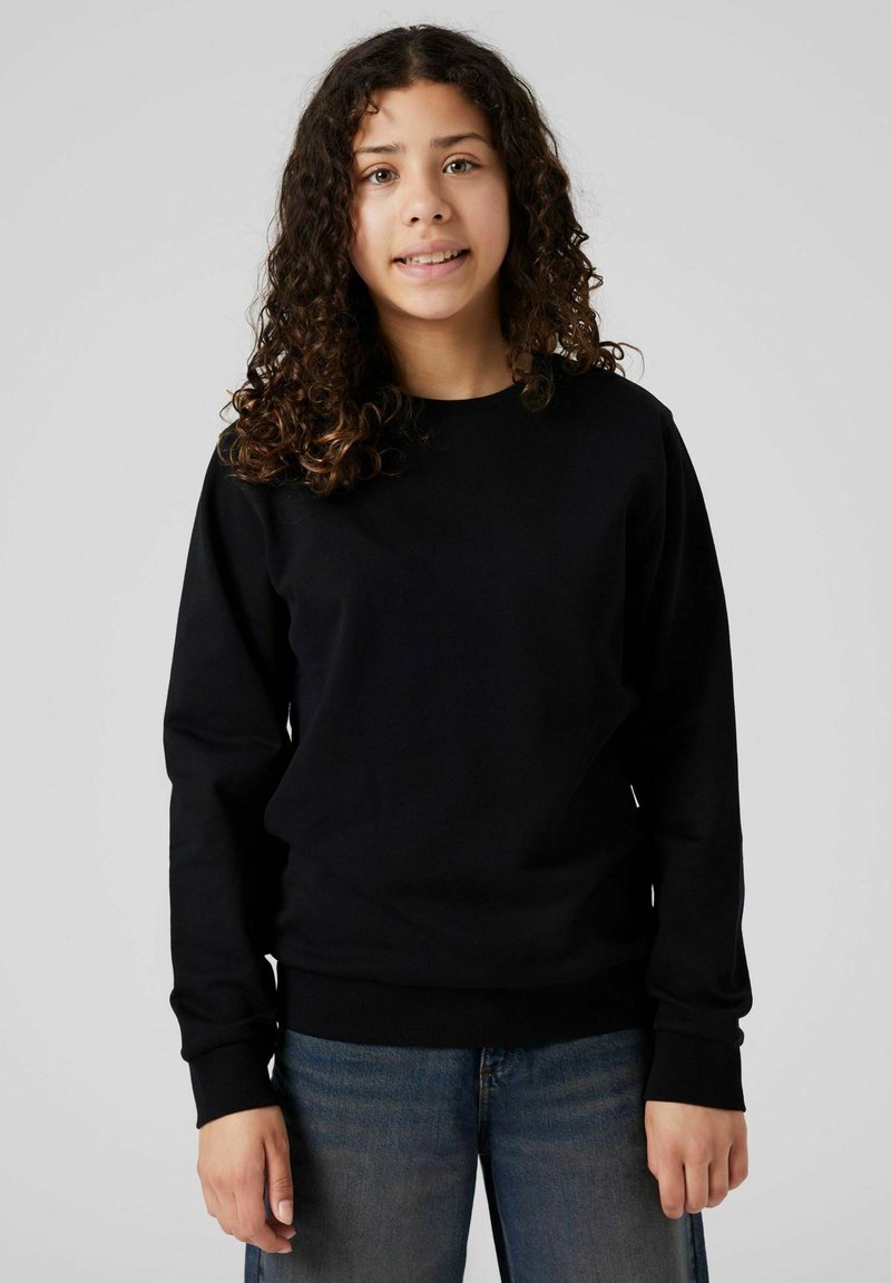 Joven con cabello rizado, vistiendo un sudadero negro y jeans azules, de pie frente a un fondo liso de color gris claro.
