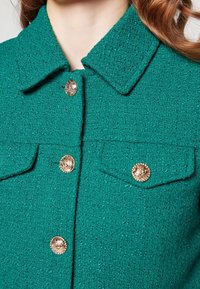 Gros plan sur une veste texturée bleu canard avec un col, deux poches à rabat et des boutons dorés ornés sur le devant et sur les poches.