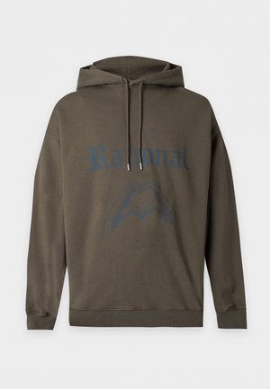 Jonge man staand met handen in de zakken, draagt een donkergroene hoodie, wit ondershirt, brede bruine broek en bruine schoenen op een witte achtergrond.