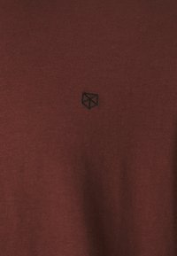 Bordo medvilninis marškinėliai su juodu geometriniu logotipu išsiuvinėtu kairėje krūtinės pusėje. Lygi tekstūra ir klasikinis siluetas.