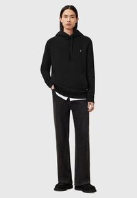 Sweat à capuche noir avec poche kangourou et cordon de serrage, associé à un jean noir ample et des chaussures slip-on tissées noires. Branding minimal visible.