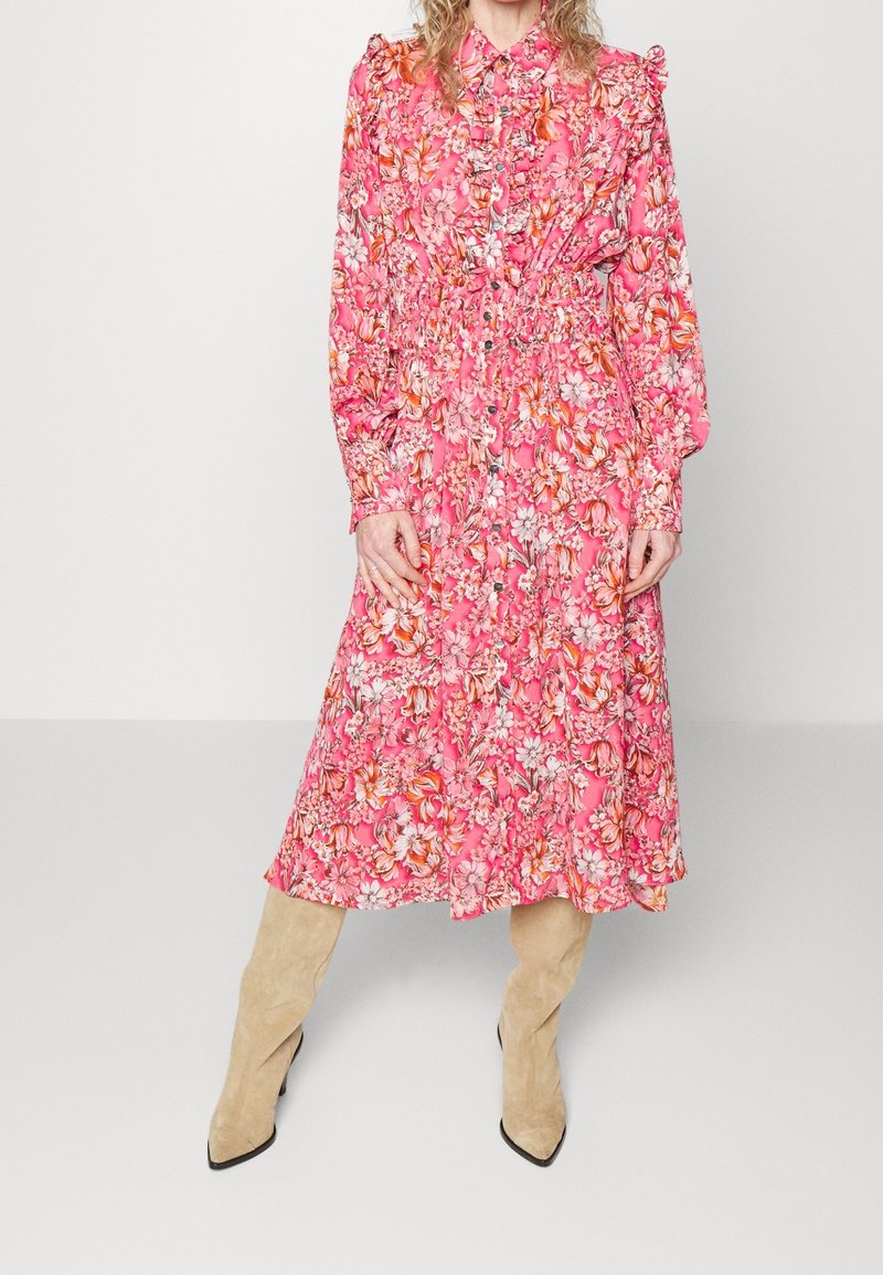 Femme portant une robe midi rose à fleurs à manches longues avec des boutons et des bottes hautes en daim beige, debout devant un fond uni.