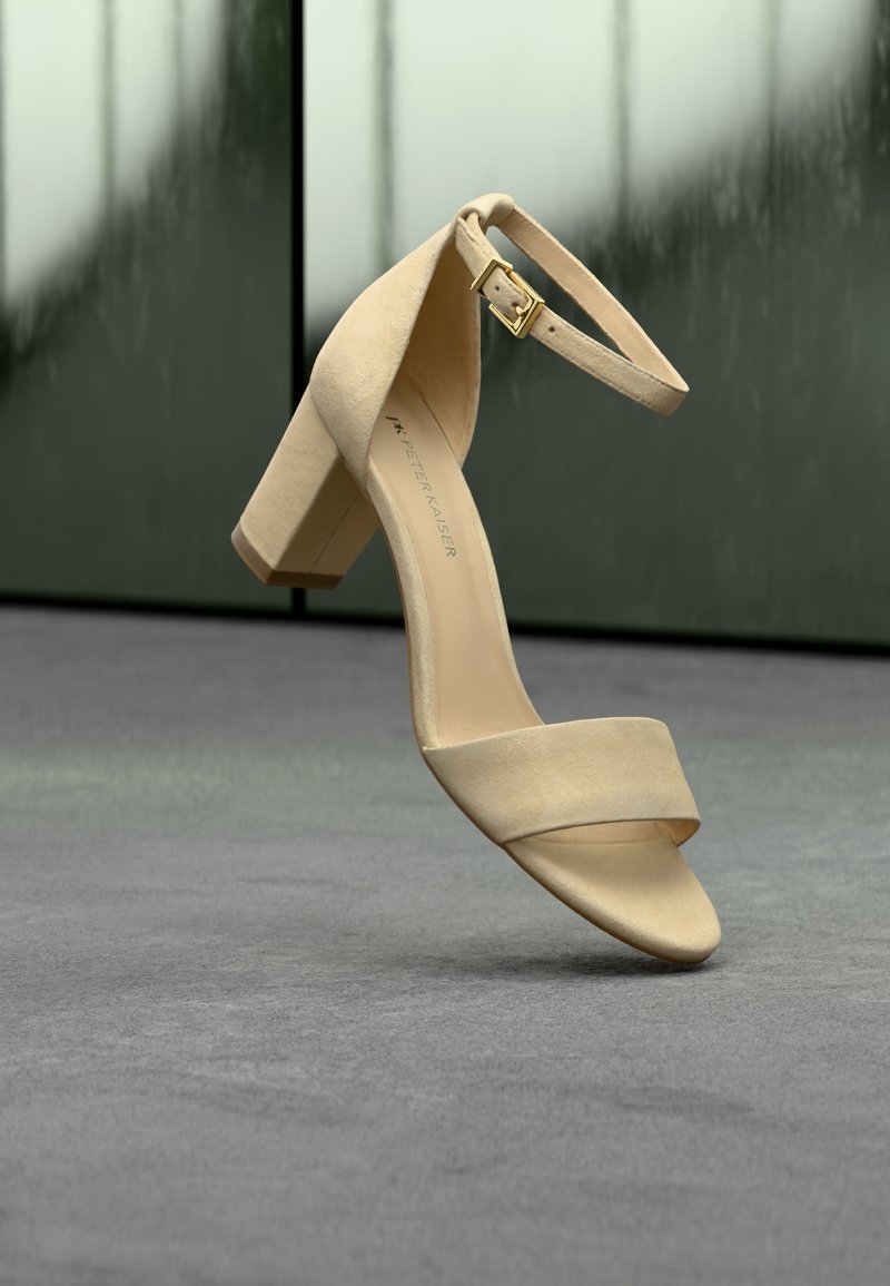 Μπεζ σανδάλι από σουέτ με τακούνι μπλοκ, λουράκι στον αστράγαλο και χρυσή αγκράφα, αιωρείται πάνω από γκρι χλοοτάπητα.