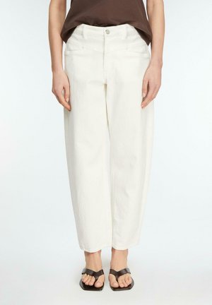 Osoba ubrana w luźne białe spodnie, brązową bluzkę i czarne sandały z odkrytymi palcami, stojąca na tle jednolitego jasnego tła.