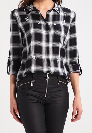 Czarna i biała koszula w kratę z kwiatową haftem, noszona z czarnymi spodniami z przednim suwakiem i kieszeniami na zamek.