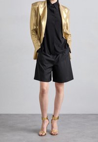 Gold metallic blazer over a black collared shirt, paired with black shorts and gold ankle strap sandals. Smooth textures and sleek design.