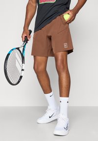 Brune idrettsshorts med sidelommer, hvit logo, kombinert med hvite sokker og joggesko, holder en tennisracket og ball.