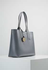 Szara skórzana torba typu tote o ustrukturyzowanym kształcie, gładkiej fakturze, z dwoma uchwytami i złotym okrągłym akcentem przymocowanym z boku.