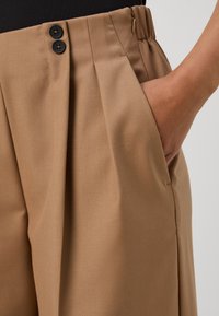 Brune plisserte bukser med to svarte knapper i livet og en sidelomme. Stoffet virker glatt med en svak glans.