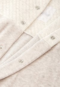 Tela acolchada de color beige claro con cuello redondeado, botones de presión plateados y textura suave. Presenta una costura de etiqueta tejida tenue.