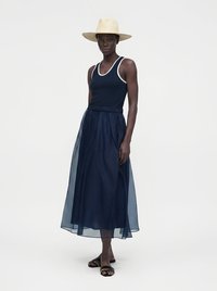 Femme portant un haut sans manches bleu marine et une jupe midi bleu marine transparente, un chapeau beige à large bord et des sandales plates noires, debout sur un fond uni.