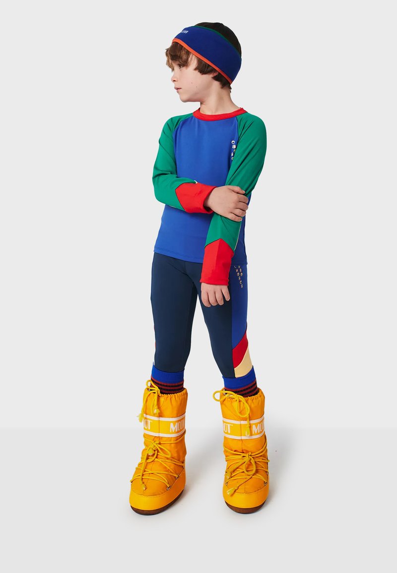 Completo da sci per bambini con parte superiore blu e dettagli verdi e rossi; pantaloni navy con pannelli multicolori; scarponi da neve giallo brillante.