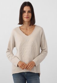Pull beige à manches longues avec un décolleté en V et un ourlet effiloché. Présente un texte en relief : « POSITIVE LIFE » et « POSITIVE MIND CLUB » avec des accents en forme de cœur. Texture douce.